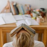 Student feeling stress and exhaustion while studying at a cluttered desk with an open book on their head.